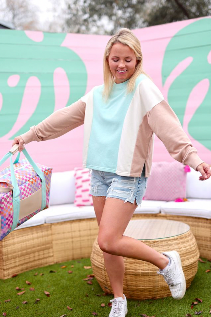 Casual two-tone sweatshirt in mint and beige, paired with distressed denim shorts and a colorful duffel bag.
