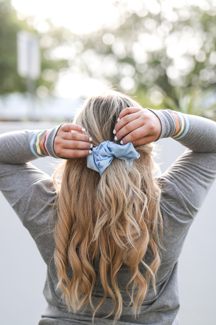 Light blue satin scrunchie styled in wavy blonde hair against a blurred outdoor background.