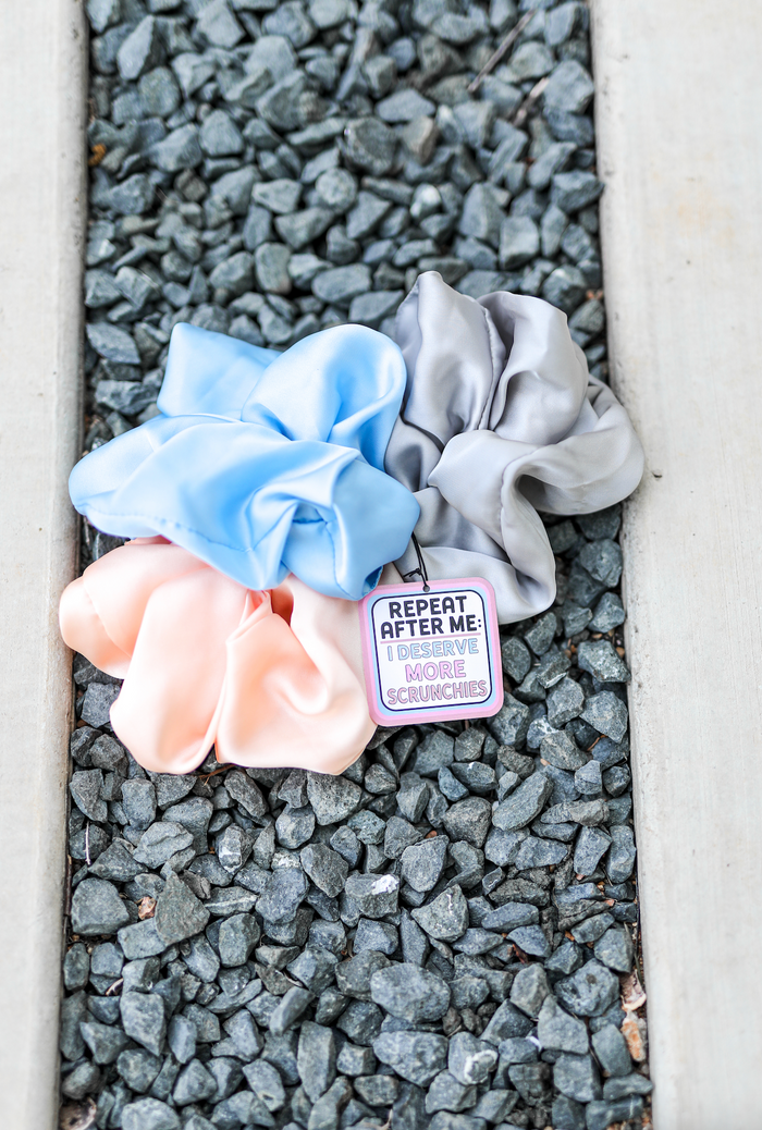 Set of satin scrunchies in blue, pink, and gray, with a playful tag saying 'I deserve more scrunchies'.