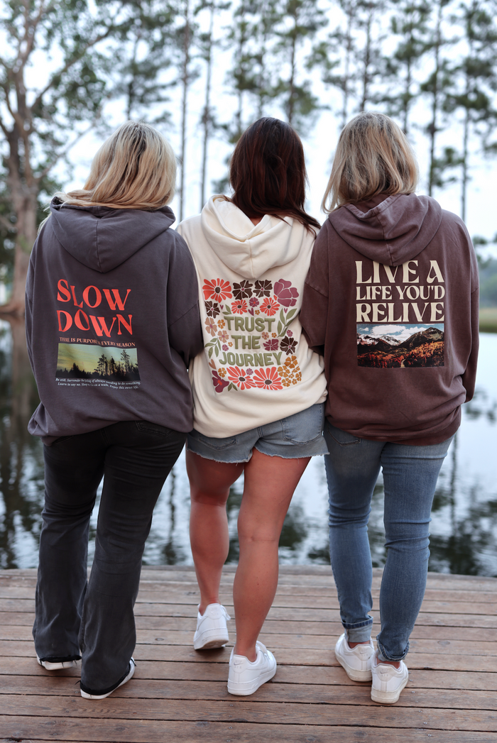 Three women in stylish hoodies: gray with 'Slow Down', cream with floral design, and brown with 'Live a Life You'd Relive'.