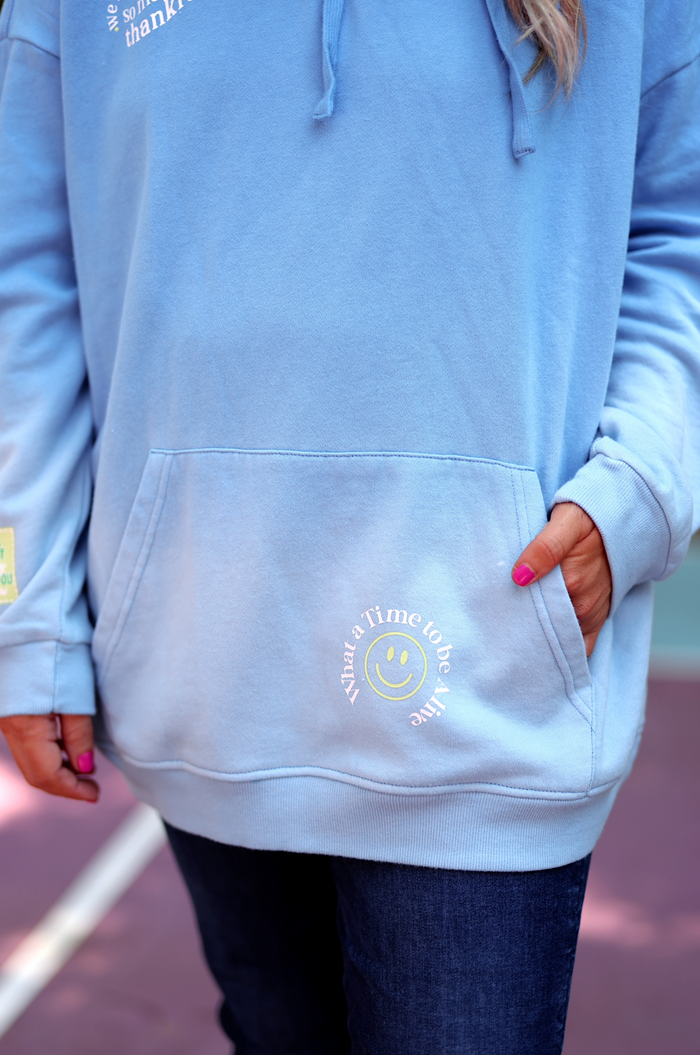 Light blue hoodie with smiley graphic, cozy material, features a front pocket and relaxed fit.