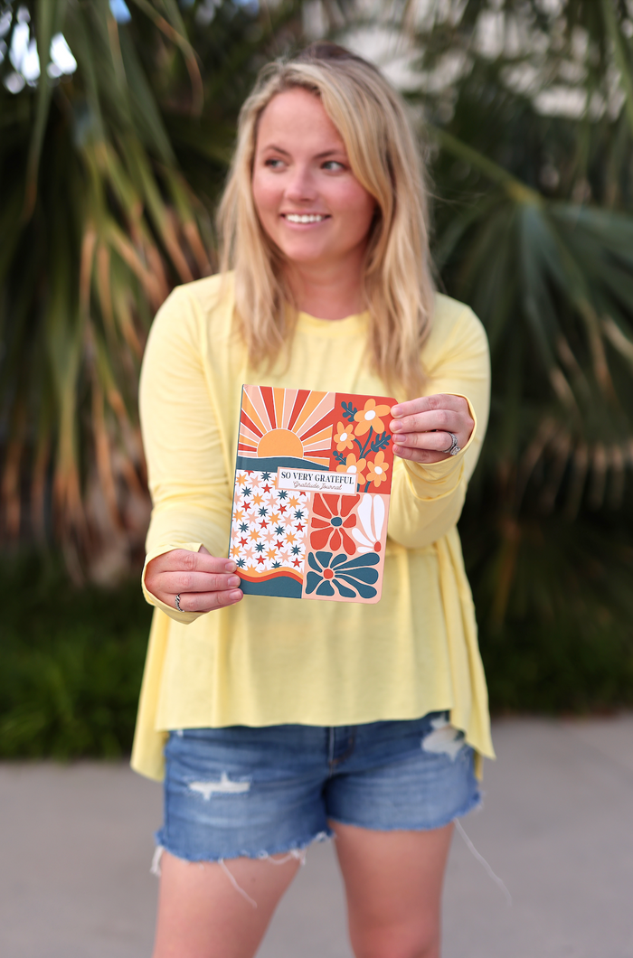 Woman holding a colorful gratitude journal with floral designs and sun motifs, wearing a yellow top and denim shorts.