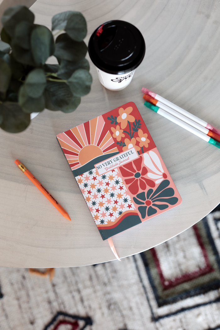Colorful gratitude journal with floral design, orange cover, surrounded by pens and coffee cup.