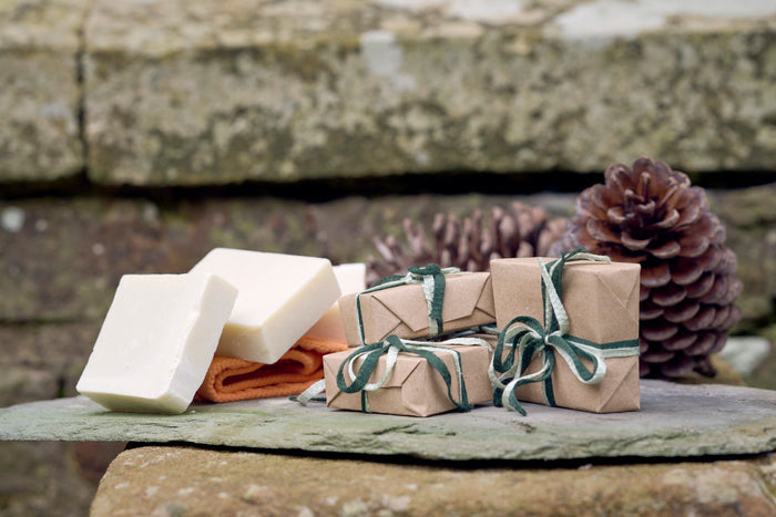 Three creamy white bars of olive oil castile soap, with three wrapped packages of soap, and a fircone in the background