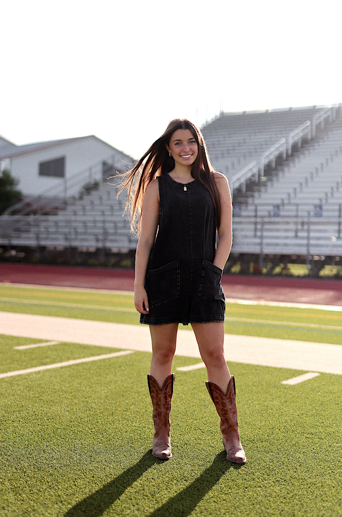 Black denim sleeveless dress with pockets, paired with brown cowboy boots on a sports field.