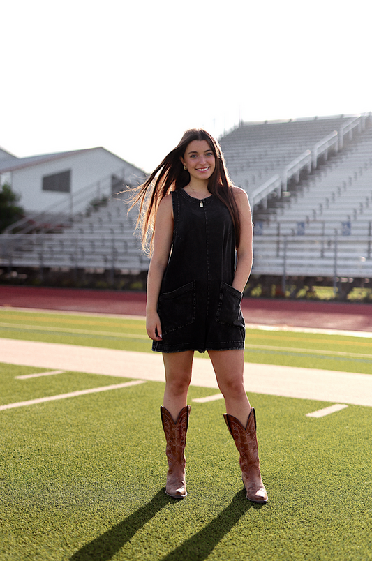 Black denim sleeveless dress with pockets, paired with brown cowboy boots on a sports field.