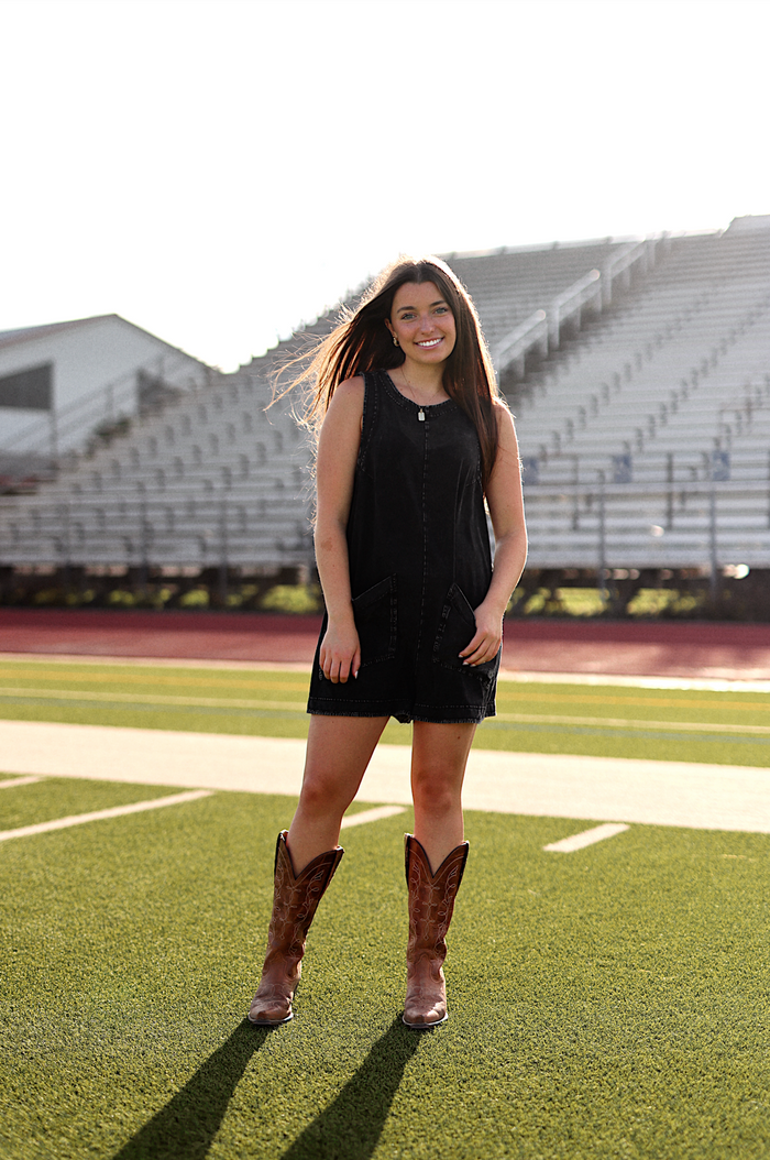 Black denim sleeveless dress with pockets, paired with brown cowboy boots on a sports field.