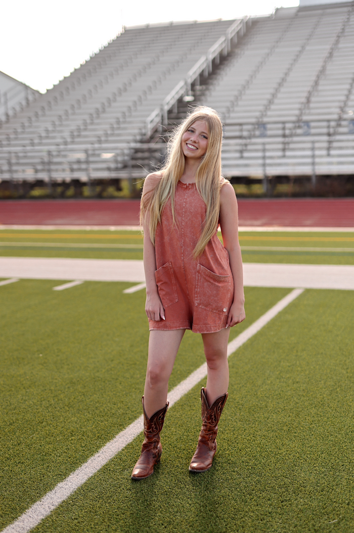 Rust orange denim romper with pockets, paired with brown cowboy boots on a sports field.