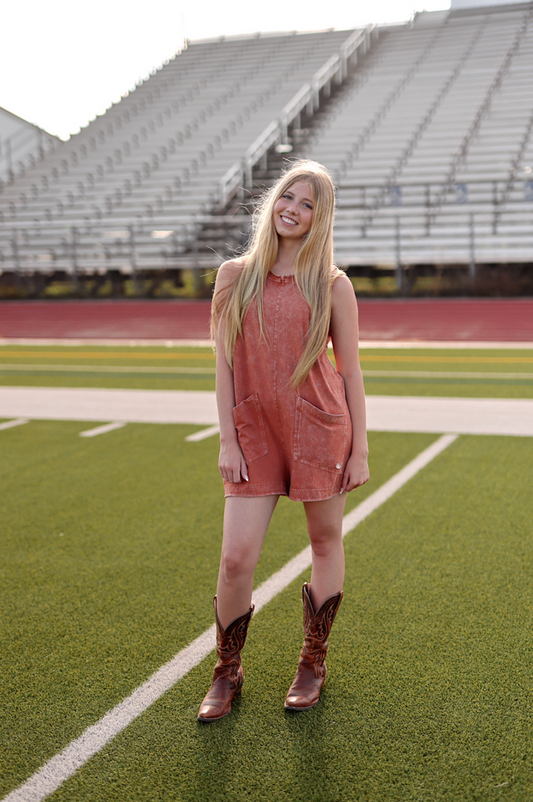 Rust orange denim romper with pockets, paired with brown cowboy boots on a sports field.