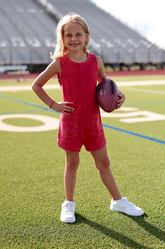 Cute girl in a red denim romper, holding a football on a grassy field, wearing white sneakers.