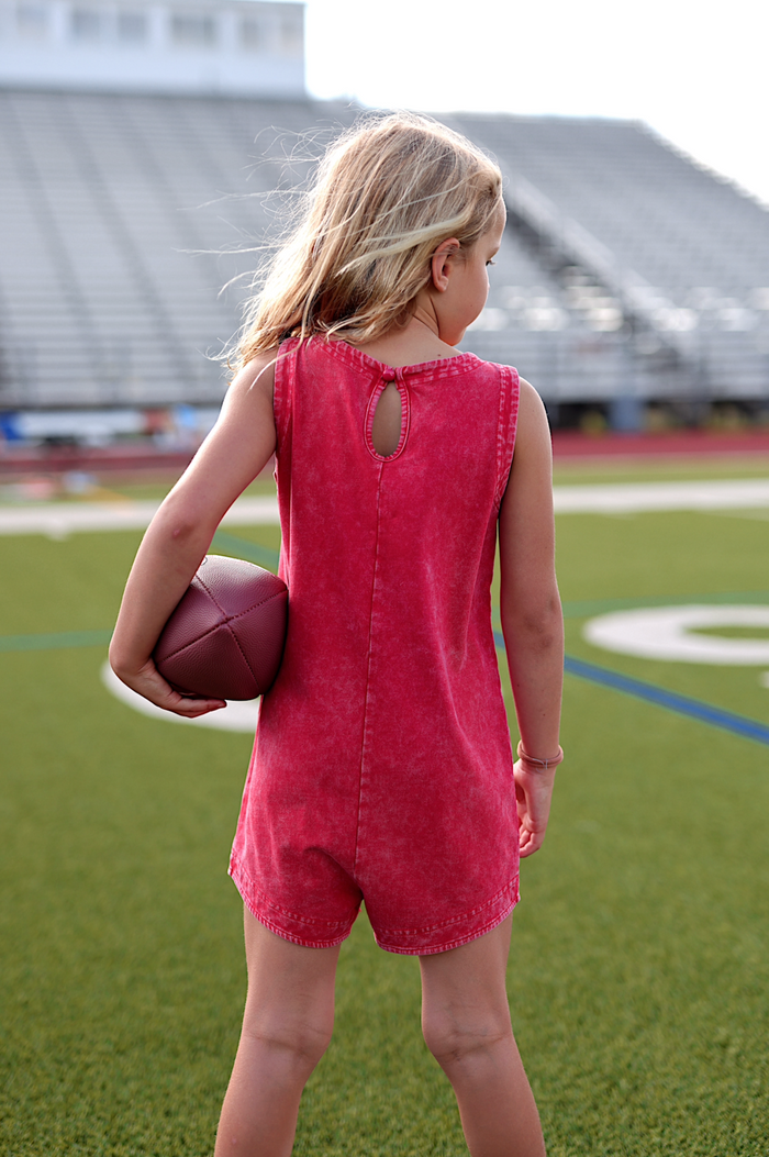 Child in a red denim playsuit holding a football, showcasing sporty summer style at a football field.