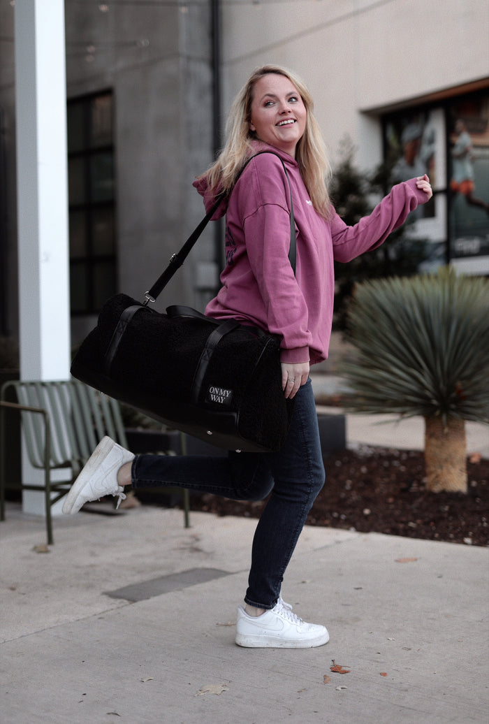 Woman in pink hoodie holding a black plush duffel bag, perfect for gym or travel.