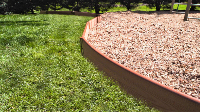 Tool-Free Classic Curved Playground Border in sienna containing wood chip safety surfacing, bordered by green grass and trees.