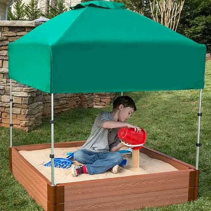 Child playing in a square composite sandbox with green telescoping canopy and sand toys outdoors