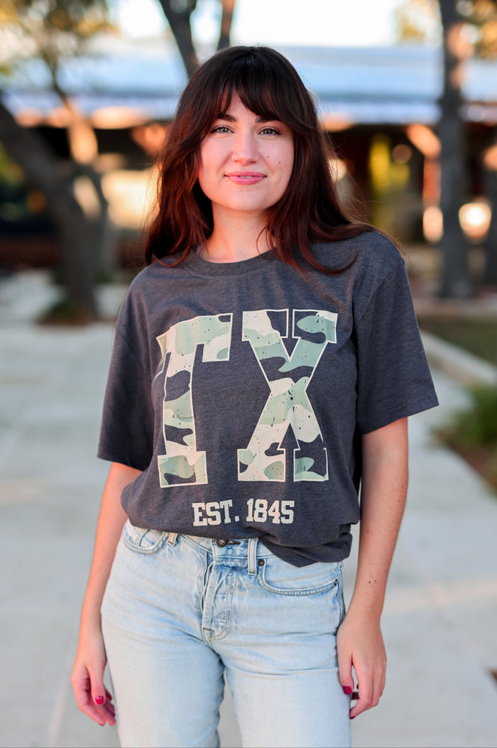 Woman wearing a gray t-shirt with 'TX' design and light blue jeans outdoors.