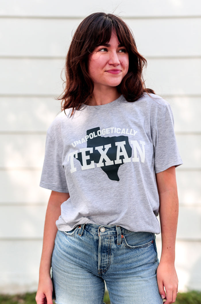 Person wearing a gray t-shirt with 'Unapologetically Texan' text and denim shorts outdoors.