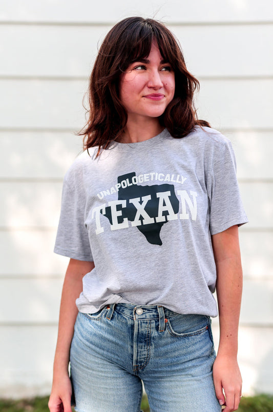 Person wearing a gray t-shirt with 'Unapologetically Texan' text and denim shorts outdoors.