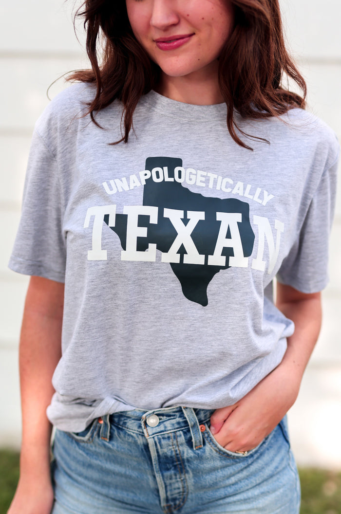 Person wearing a gray t-shirt with 'Unapologetically Texas' design and denim shorts outdoors.