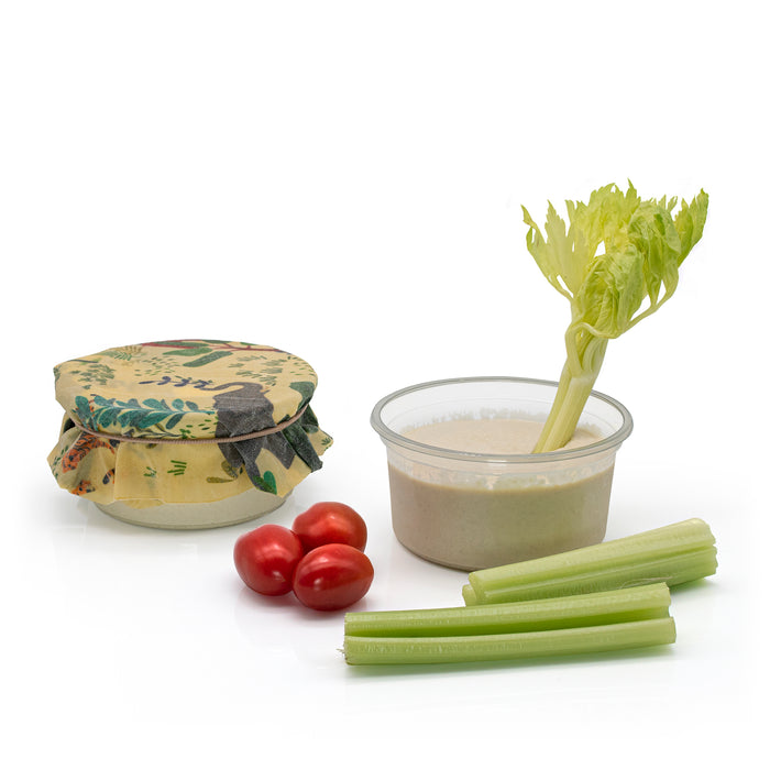 A set of reusable food storage items on a white background, including a small glass jar with a floral-patterned fabric lid, a clear plastic cup filled with dip and topped with a celery stalk, and three celery sticks with two cherry tomatoes.
