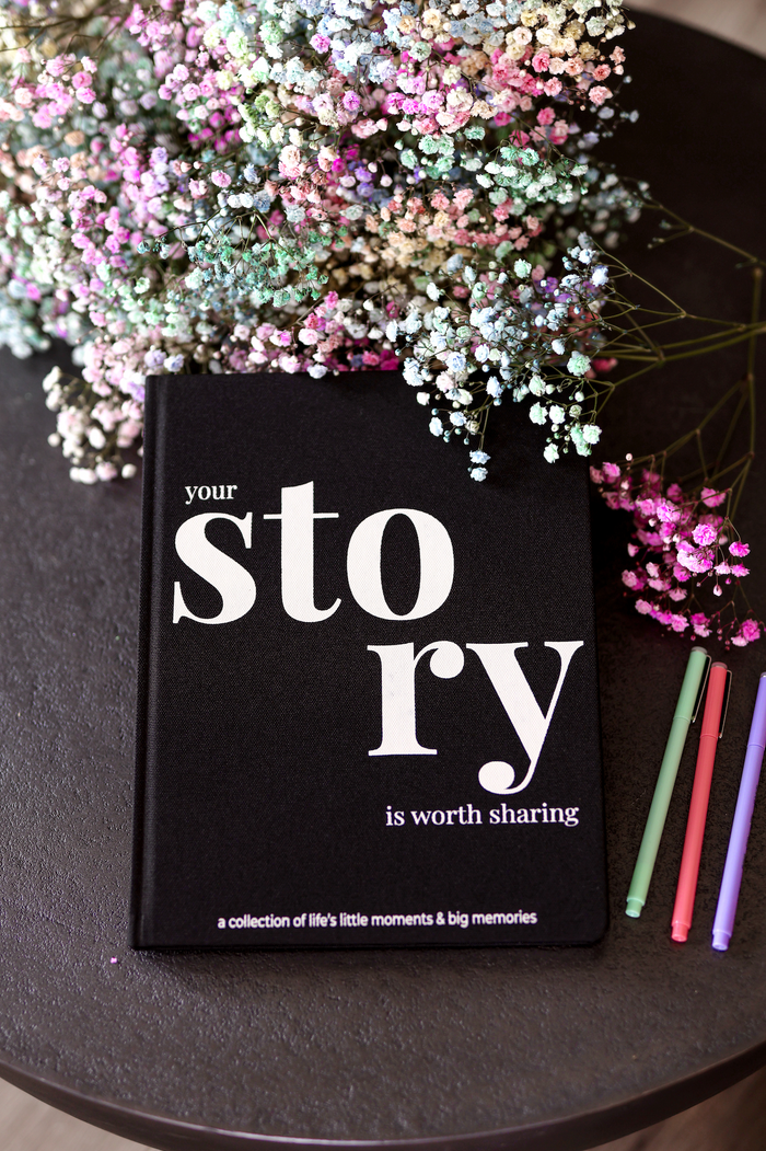 Black hardcover journal titled 'Your Story Is Worth Sharing' with colorful pens and floral background.