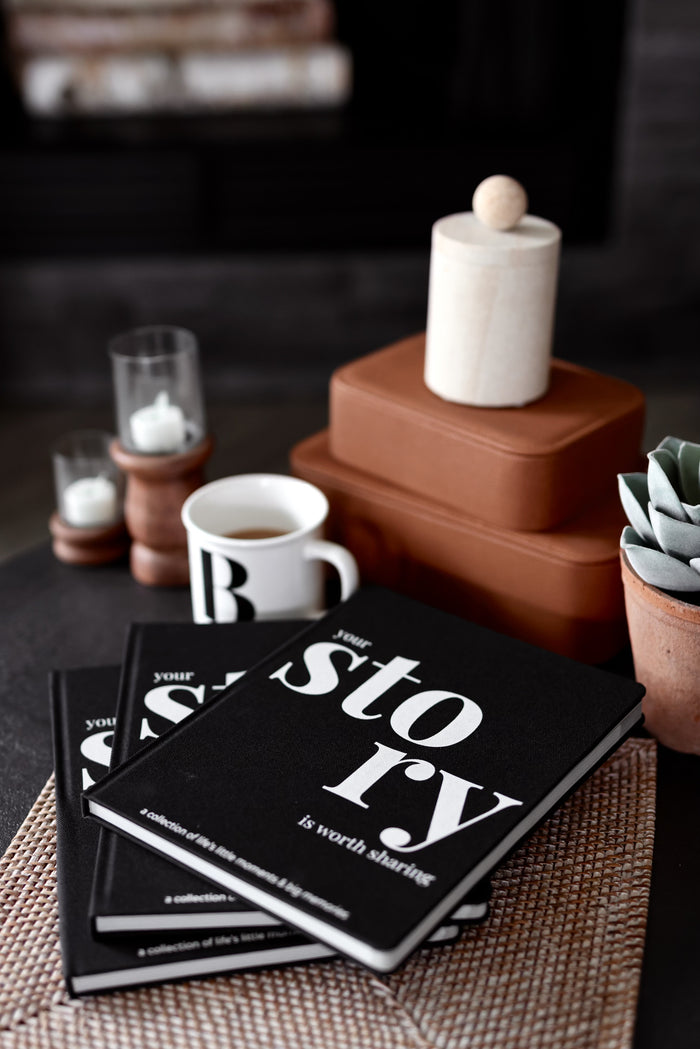 Black hardcover journal 'your story is worth sharing' with coffee mug and decorative items on a table.
