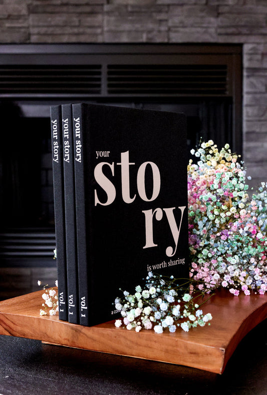 Set of three black storybooks with floral accents on a wooden display, designed for sharing personal stories.