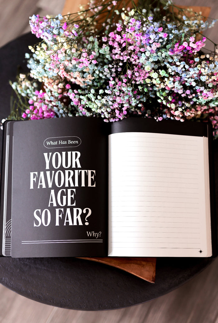 Open black journal with prompts about favorite age and blank pages, surrounded by colorful dried flowers.